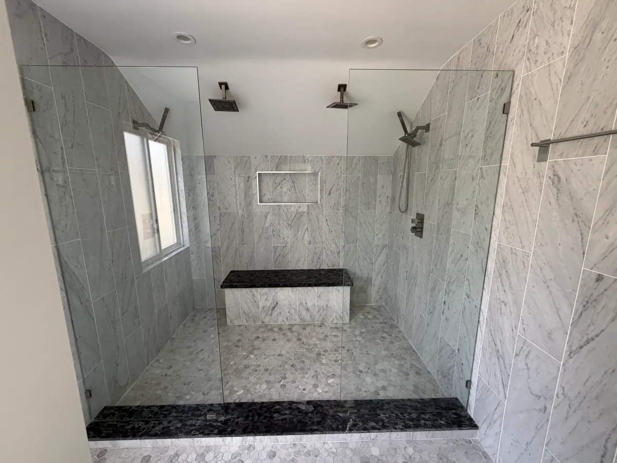 Large walk-in shower with two ceiling-mount showerheads, a handheld wand, marble-look wall tile, a built-in bench and frameless glass