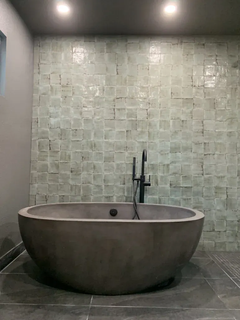 Freestanding stone soaking tub with a floor-mounted black faucet against a handmade green tile wall
