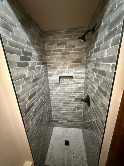 Walk-in shower with gray glazed wall tile, a recessed niche, matte black fixtures and a white hexagon mosaic floor