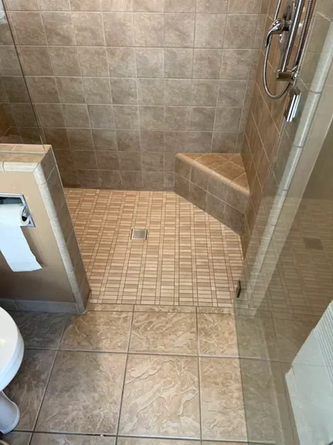 Walk-in shower with tan tile, a corner bench, a low curb and a handheld shower on a slide bar
