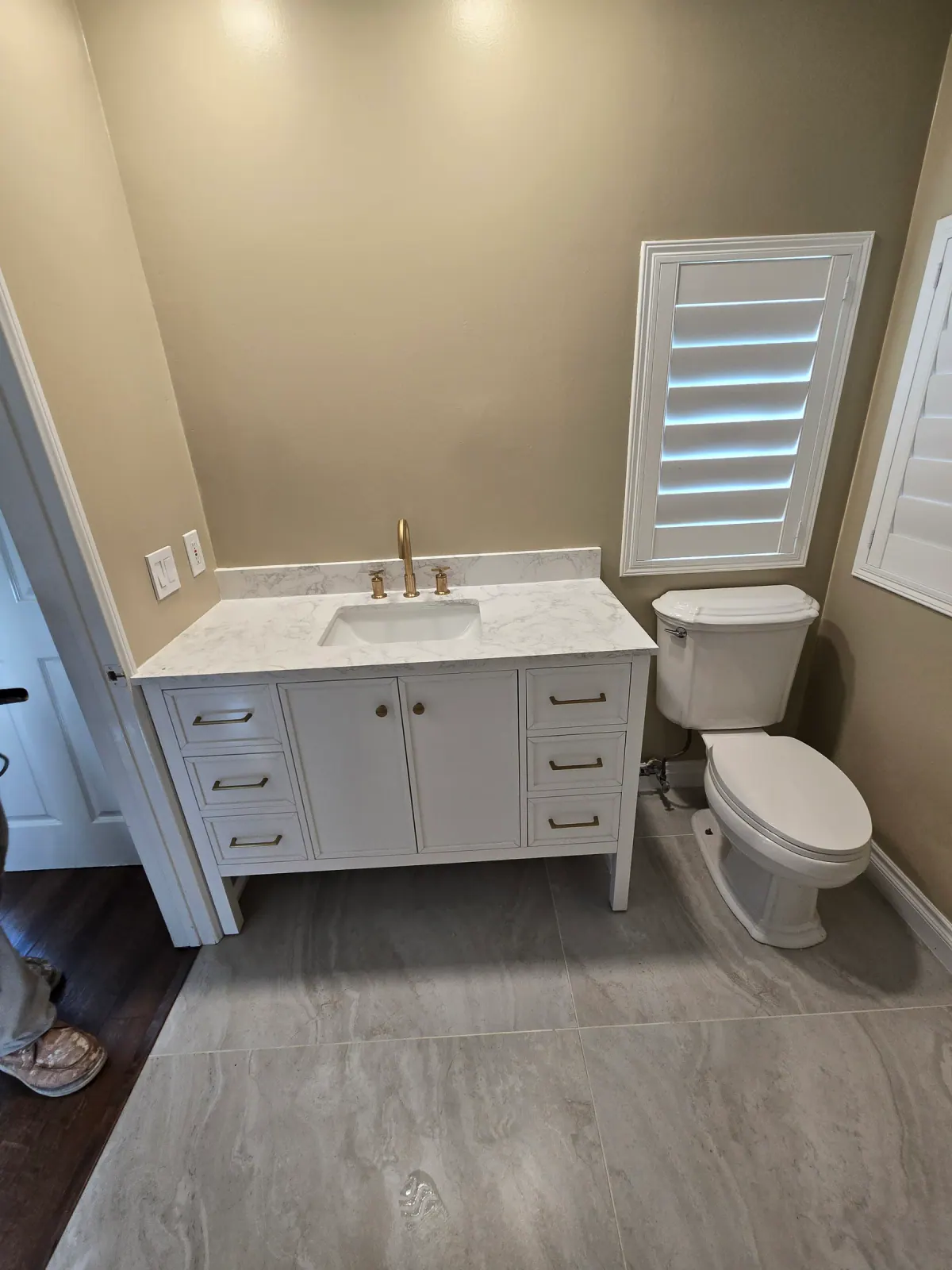 Bathroom with a white vanity, white stone top, brass faucet, new toilet and large gray floor tile beside a shuttered window