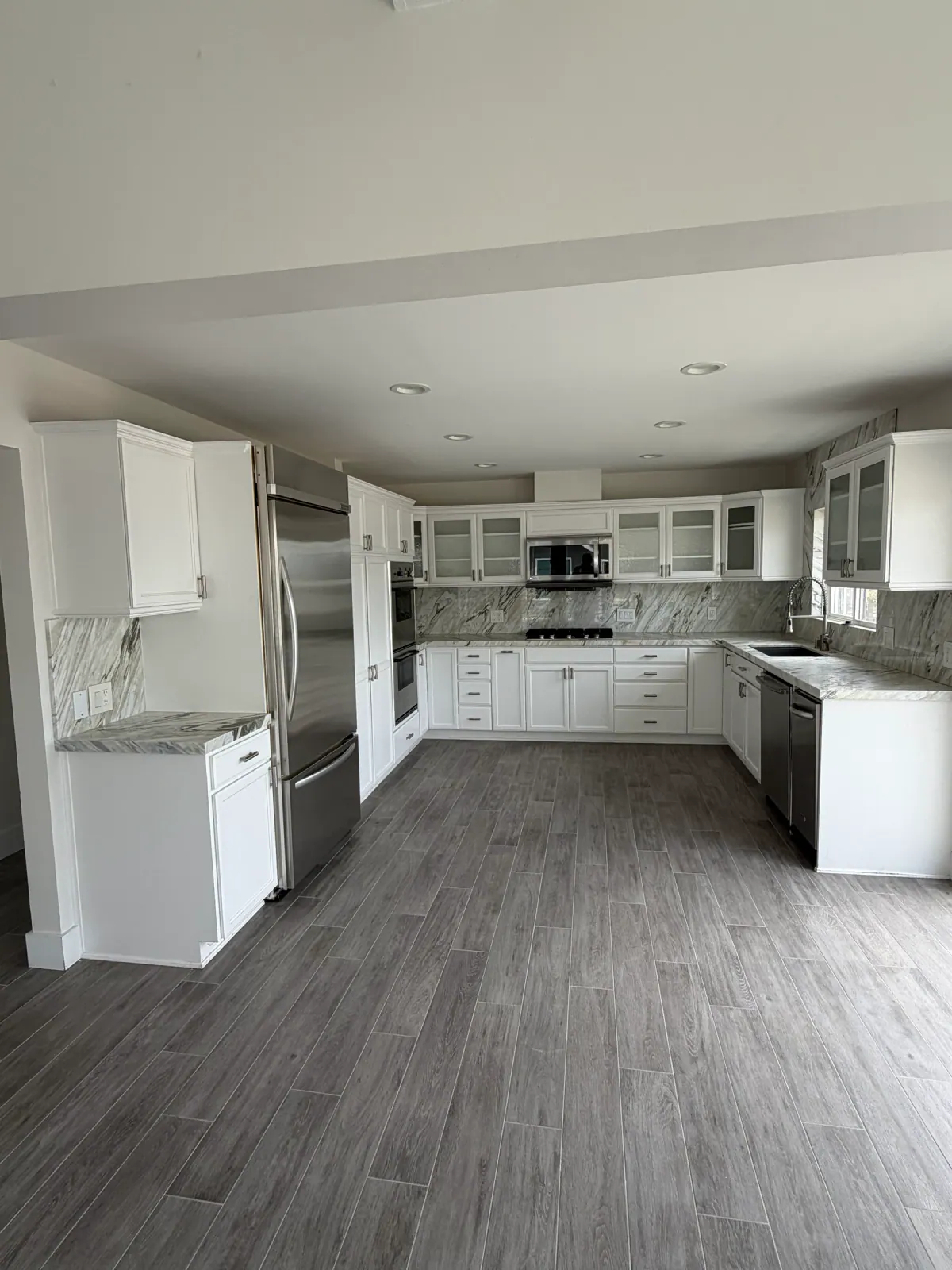 Remodeled U-shaped kitchen with white shaker cabinets, glass-front uppers, stainless appliances, veined stone counters and backsplash and wood-look plank tile