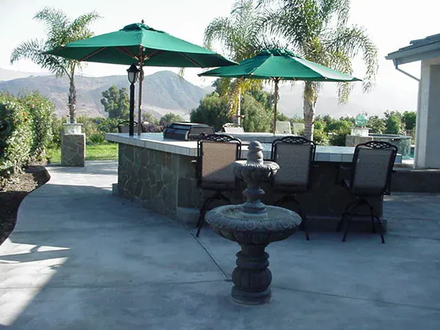 Outdoor kitchen and bar with stone-faced counter, green patio umbrellas and a tiered fountain