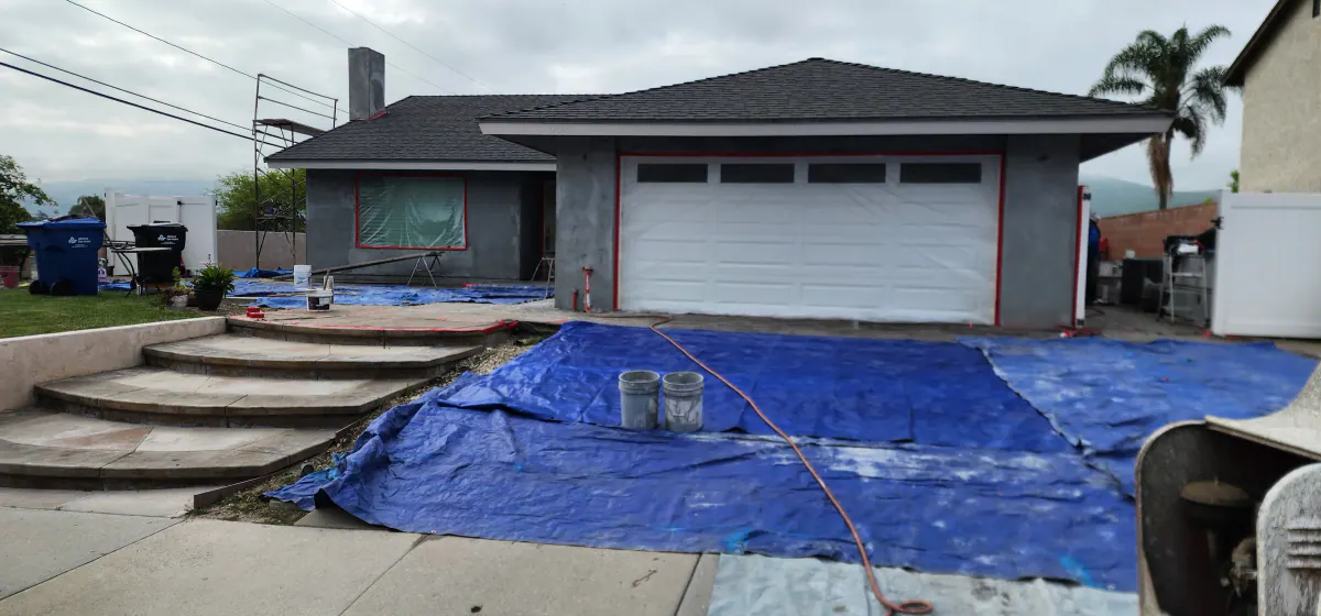 Front of a single-story house in gray base-coat stucco during a re-stucco, with scaffolding at the side and blue tarps covering the entry steps