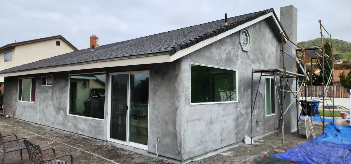 Back of a single-story house in gray base-coat stucco with a sliding glass door, new window openings and scaffolding at the corner