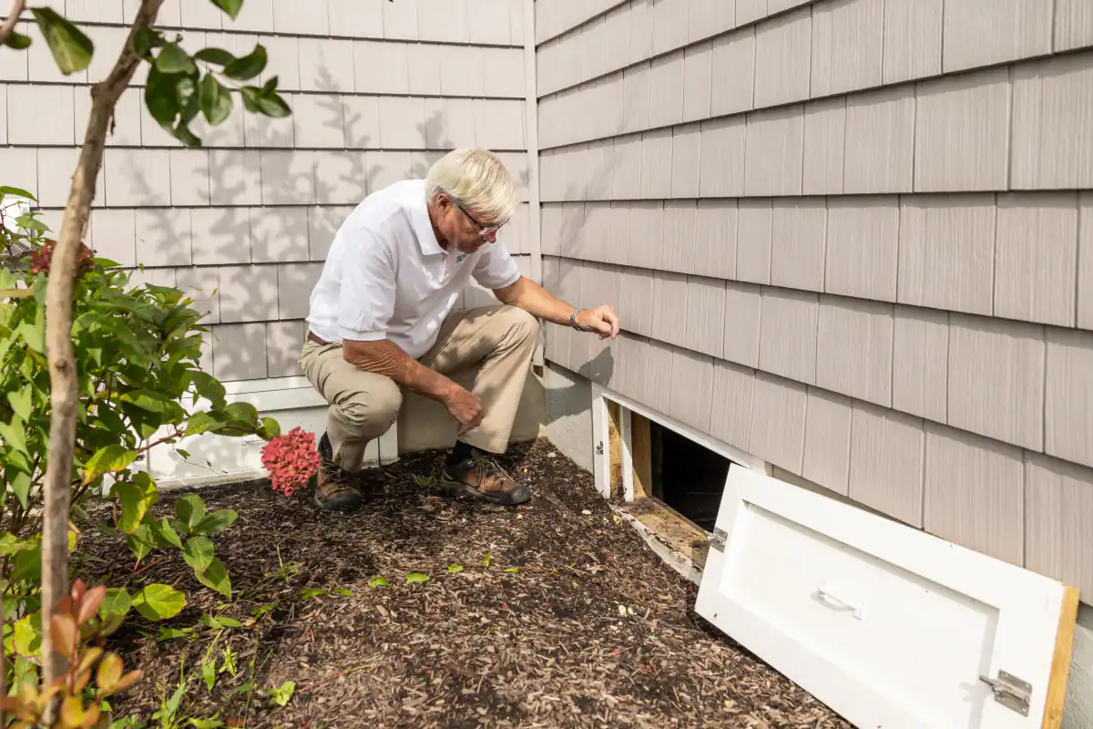Inspector crouching beside an open crawl space access door at the base of a house with gray siding